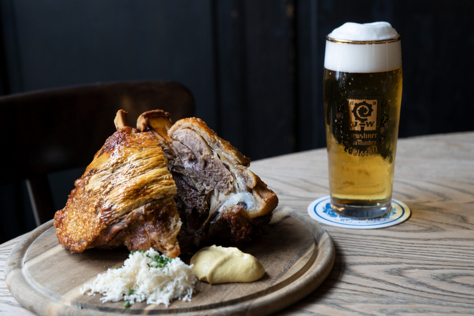 Eine rustikale Fleischspezialität wird auf einem Holzteller neben einem frisch gezapften Bier serviert.