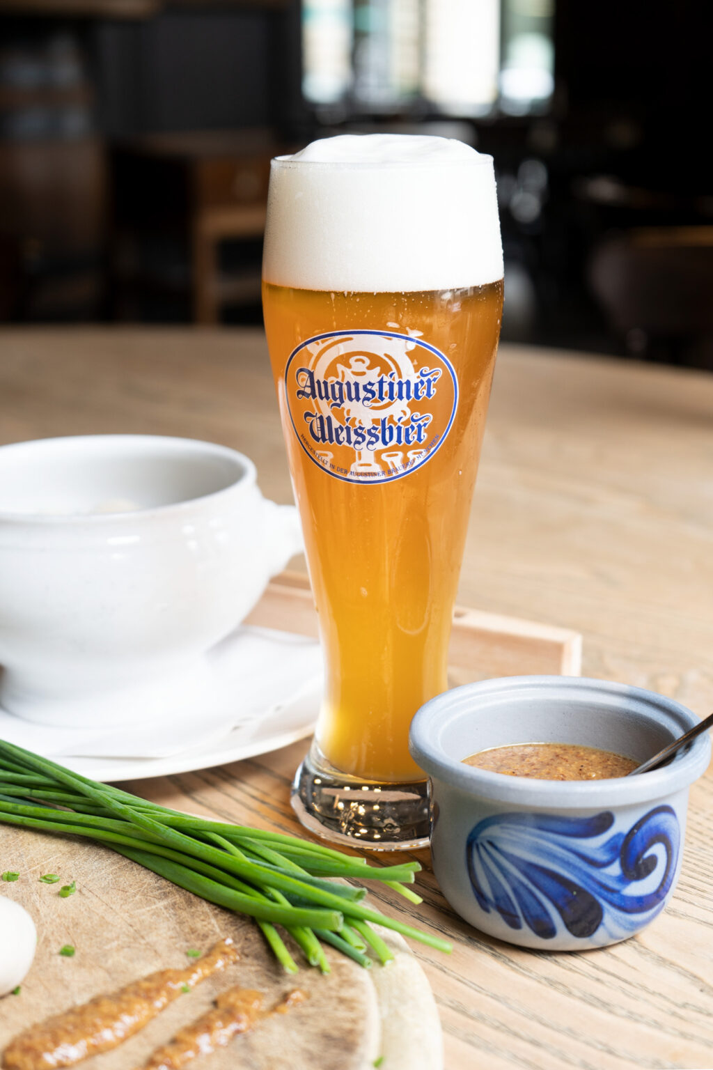 Frühstücksähnliches Arrangement mit Weißwürsten, Brezel, Schnittlauch, Weizenbier und Kaffee auf hellem Holztisch.