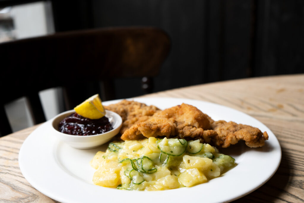 Paniertes Schnitzel mit Kartoffel-Gurken-Salat, Preiselbeeren und Zitronenspalte auf einem weißen Teller.