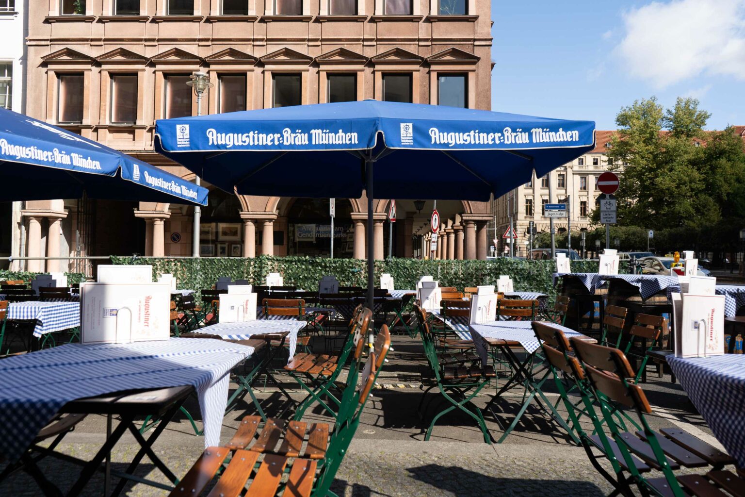 Biergarten-Terrasse des Augustiner am Gendarmenmarkt mit gedeckten Tischen, Holzstühlen und blauen Augustiner-Bräu-Sonnenschirmen vor historischer Stadtkulisse.