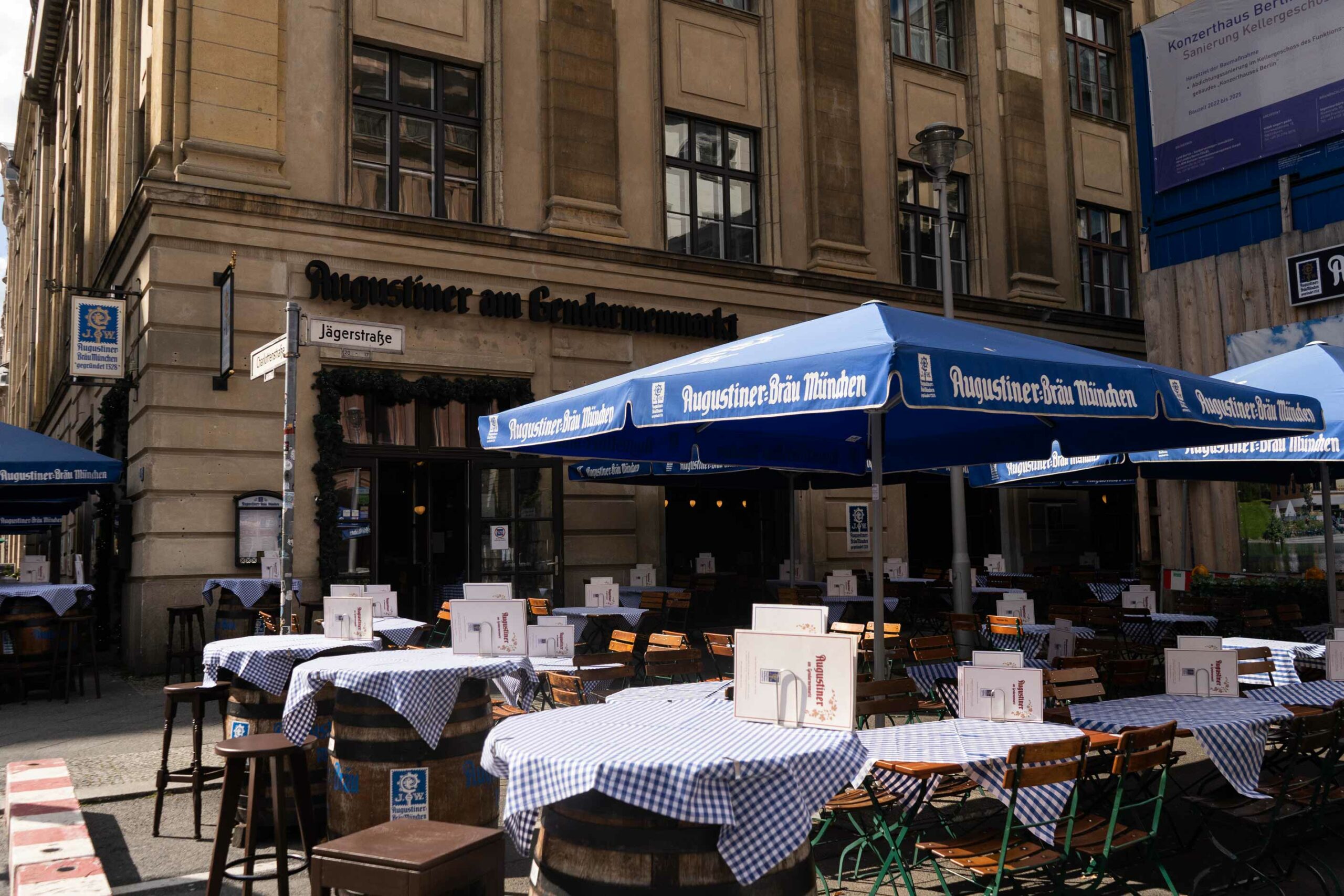 Biergarten-Terrasse vor dem Augustiner mit mehreren gedeckten Tischen, Holzstühlen und großen blauen Augustiner-Bräu-Sonnenschirmen.